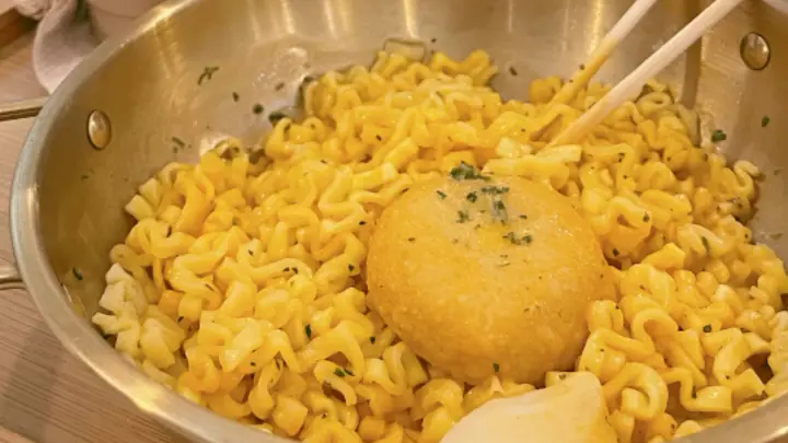 A gold DIY cooking pot of cheesy ramyeon topped with a fried patty, cooked at the Ramyeon Bar